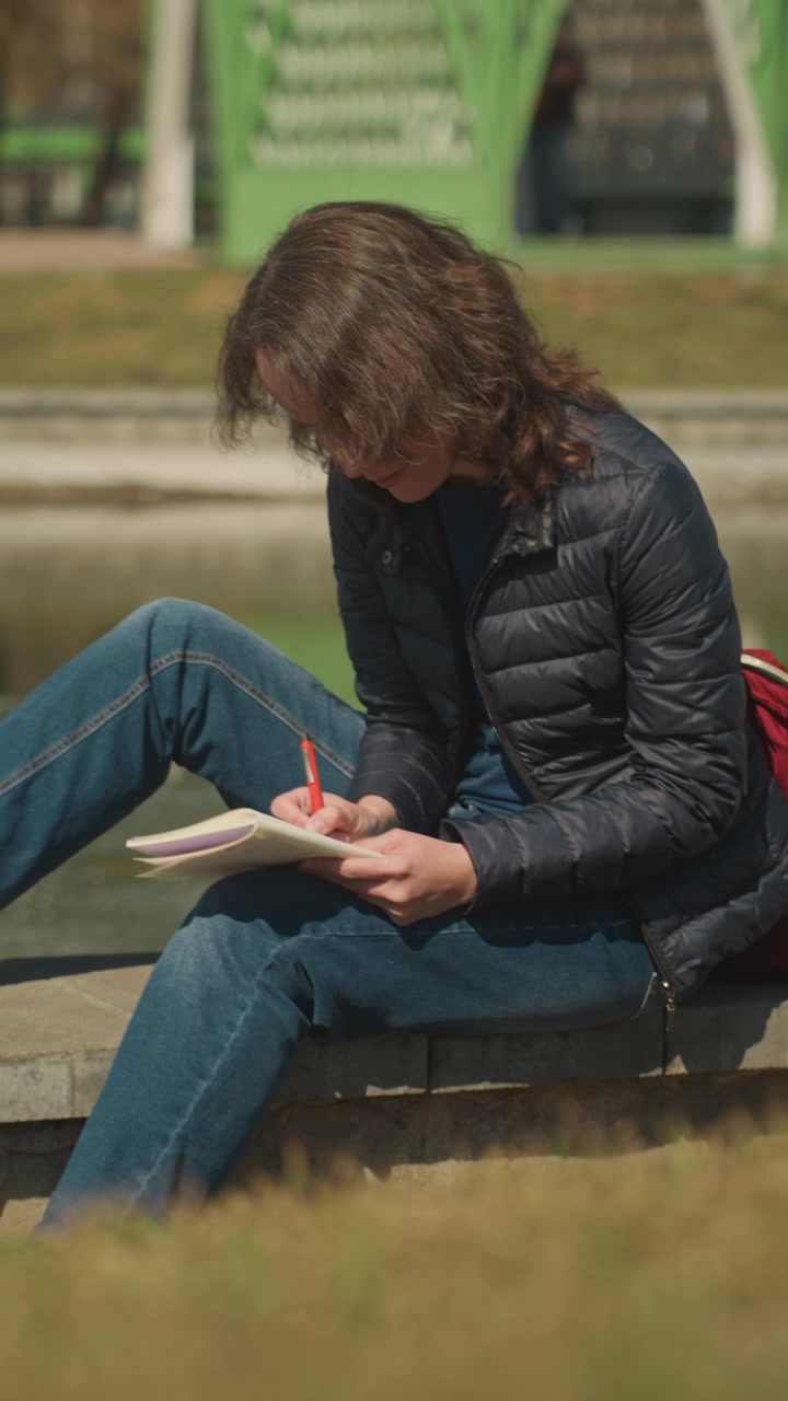 Close up of woman writing in notebook with red pen while sitting outdoors on stone ledge. Notebook and hand visible background shows green structure and grassy area. Soft natural light