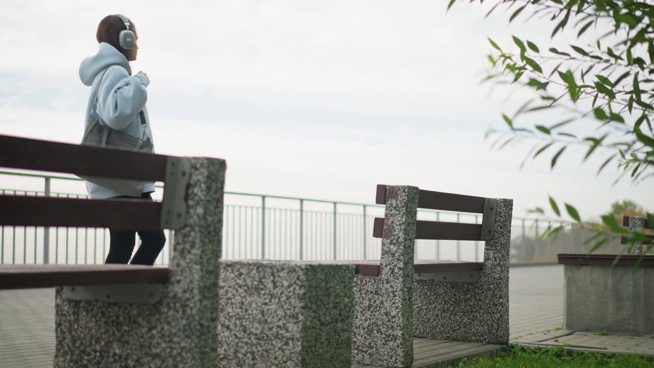 Back view of young woman walking toward concrete bench to sit, carrying bag in peaceful park setting, with green leaves and railing in background, perfect for leisure, walking, and nature shots