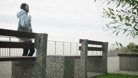 back view of young woman walking toward concrete bench to sit, carrying bag in peaceful park setting, with green leaves and railing in background, perfect for leisure, walking, and nature shots