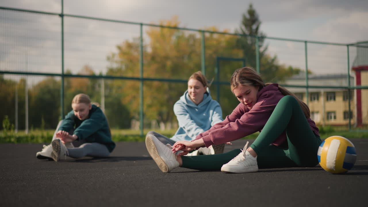 Ladies working out, seated on the ground stretching with volleyball rolling beside them, outdoor setting with iron fence, trees, and a building in the background