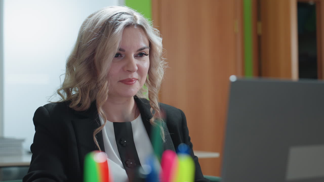 Close up of excited woman in formal attire getting set for work, smiling while interacting with laptop, foreground shows colorful pens in holder, background includes green wall