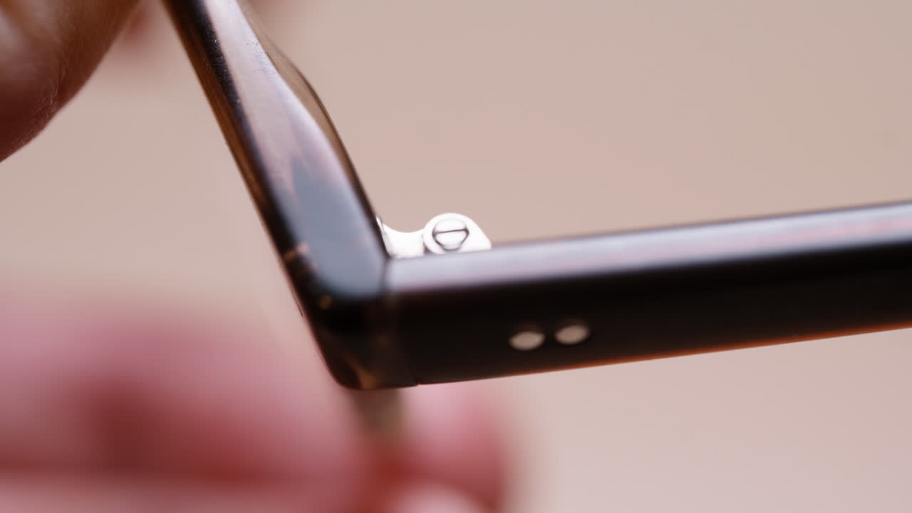 Macro shot of hands holding damaged eyeglasses up close, carefully studying the broken hinge. Fine frame details and wear clearly visible. Real and honest DIY inspection moment