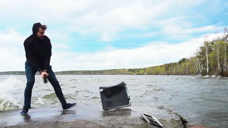 man destroying a computer by hitting it with a hammer on the shore of a lake