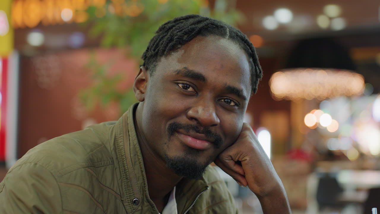 Close up of man posing with subtle smile resting hand under chin, wearing casual jacket in indoor setting with blurred bokeh lights, confident thoughtful expression
