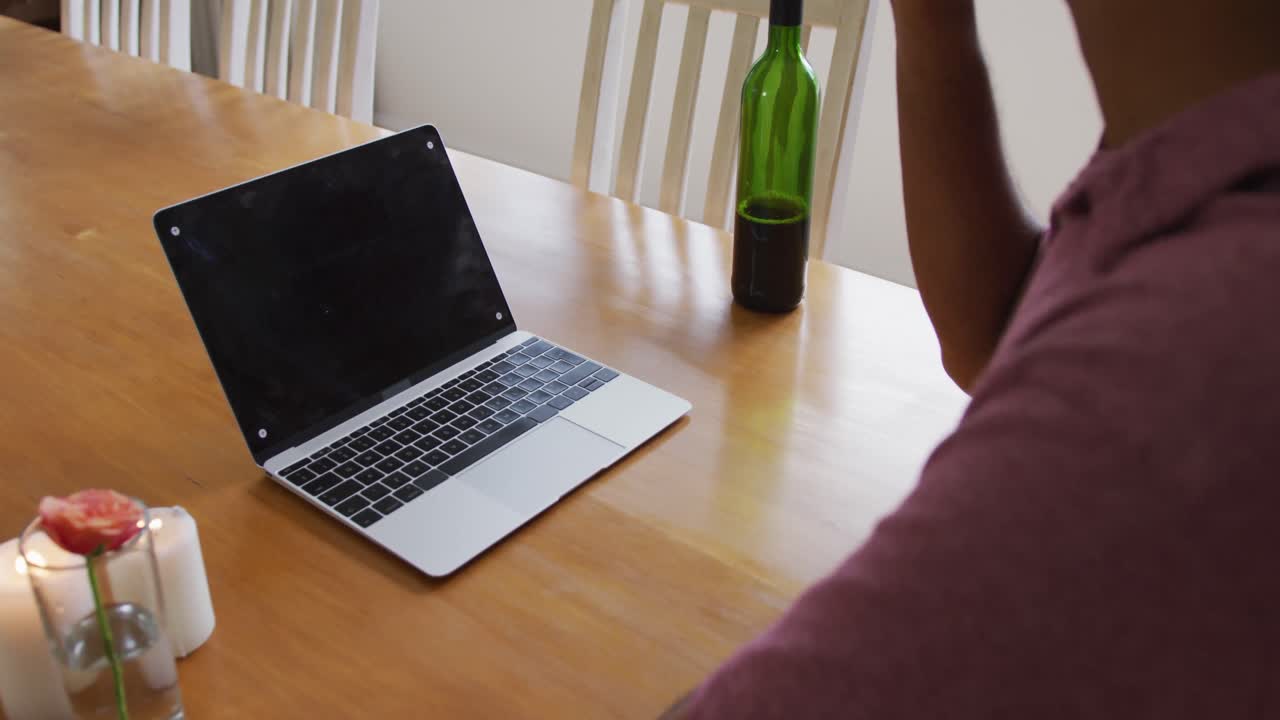 Midsection of mixed race man sitting at table using laptop drinking red wine