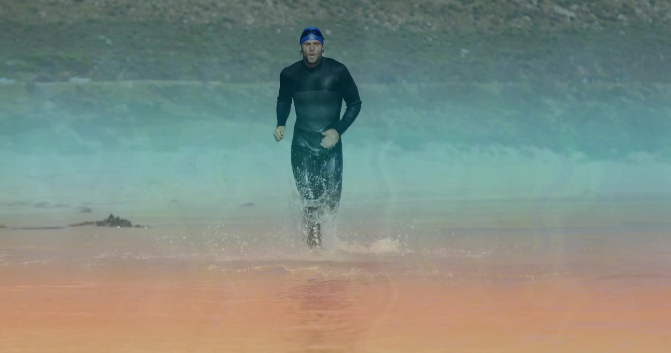Muž v černém neoprenu a modré plavecké čepici rozrážející vodní hladinu, běžící mělčinou pro triatlon.