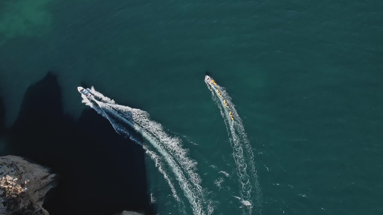 Aerial drone steadily moves forward parallel to a boat and group of kayaks, both cutting smooth wakes across the turquoise waters and casting dramatic shadows near the sheer cliffs of Ponta da Piedade