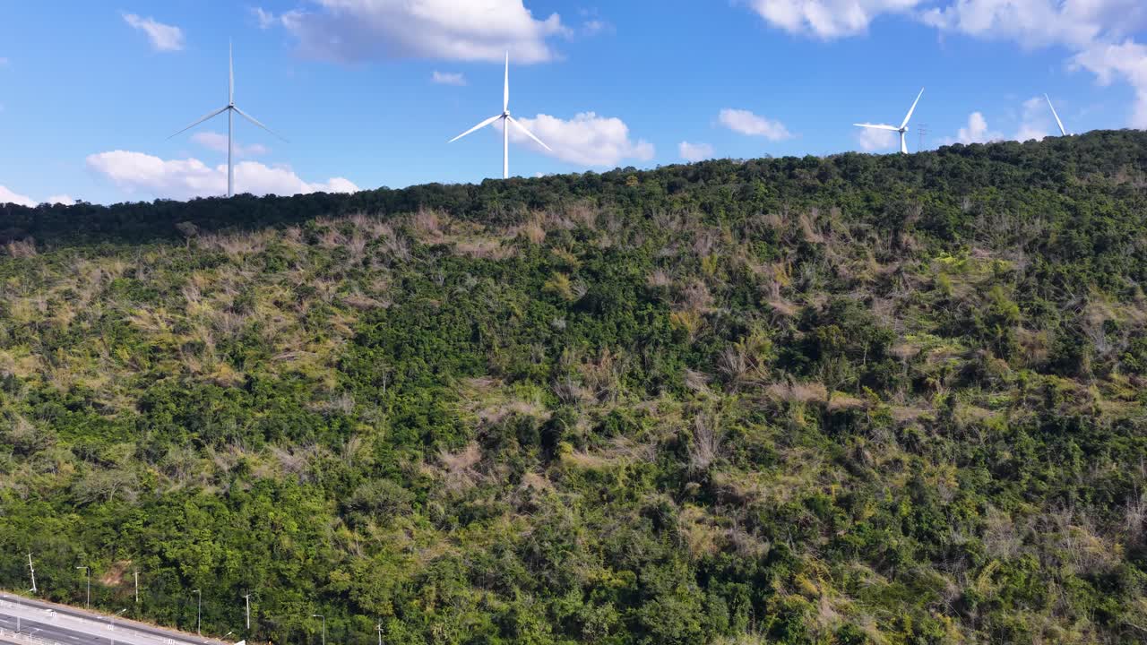 Plano aéreo amplo de uma rodovia elevada passando por uma montanha arborizada com turbinas eólicas girando