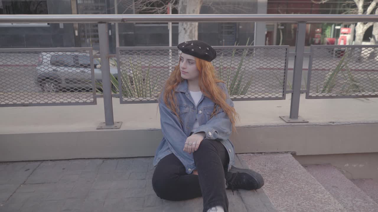 Two friends have fun taking pictures in the street. The redhead woman model poses sitting on the sidewalk for her photographer friend and smile