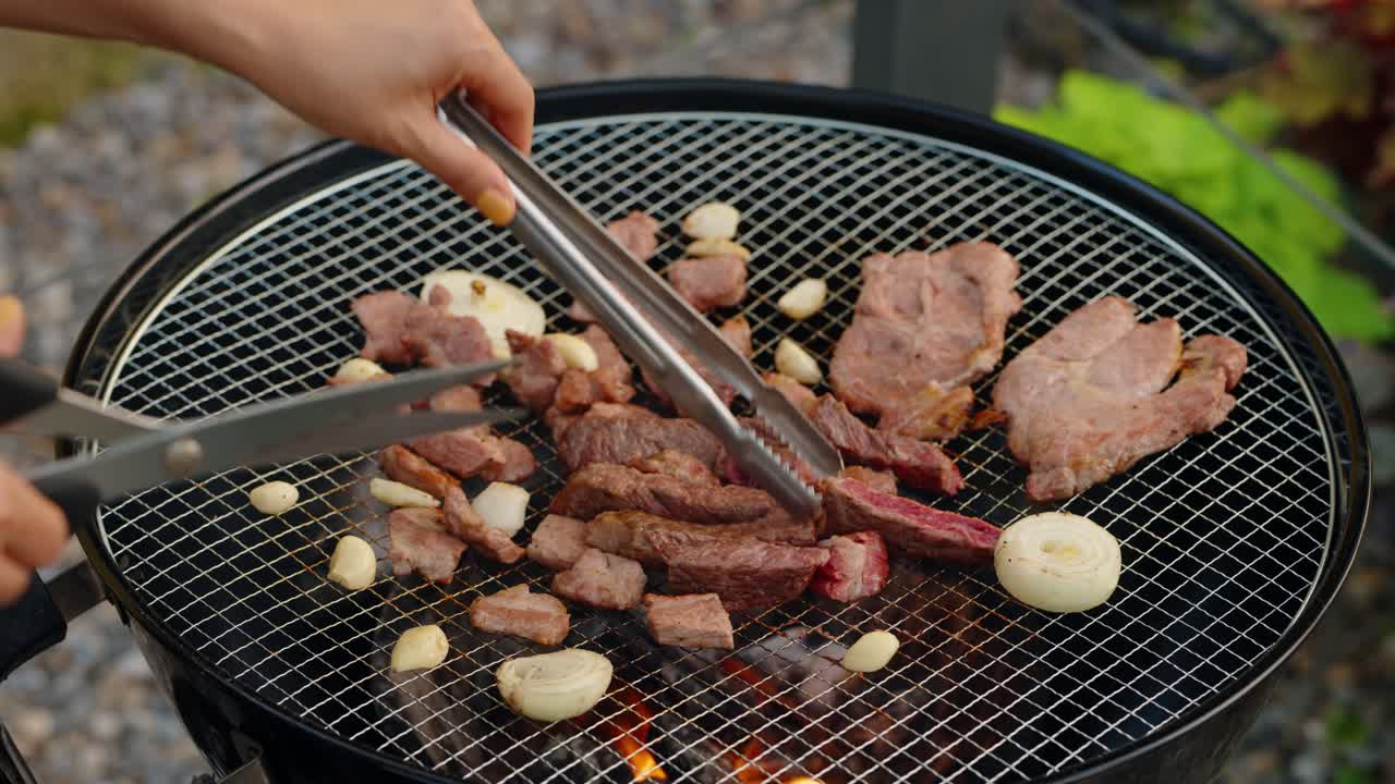 Close-up of a person flipping cooked, bite-sized beef pieces with tongs while holding scissors, grilling with garlic and onions on a hot charcoal Korean-style barbecue