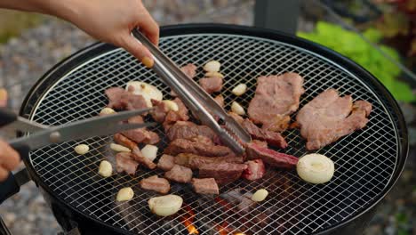 close-up of a person flipping cooked, bite-sized beef pieces with tongs while holding scissors, grilling with garlic and onions on a hot charcoal korean-style barbecue