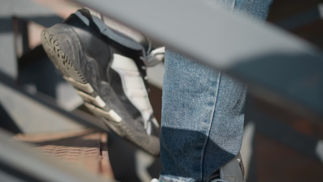 close up leg view of person wearing faded blue jeans and black white canvas sneakers descending outdoor metal staircase with brick wall backdrop sunlight casting shadows on steps and denim hem