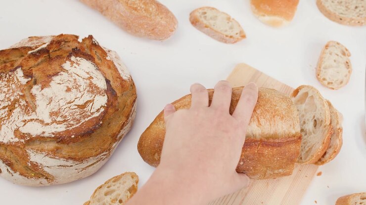 Overhead view of hands using a knife to slice crusty sourdough on a white surface.