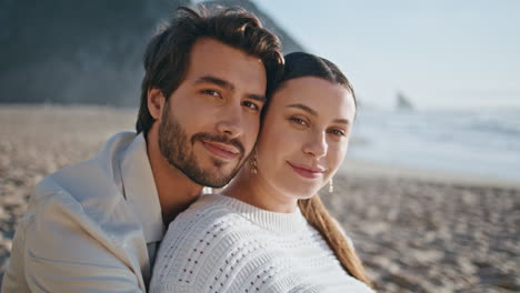 porträt von einem romantischen paar auf dem strand, das mit einem lächeln in die kamera schaut. ehegatten sitzen an der küste