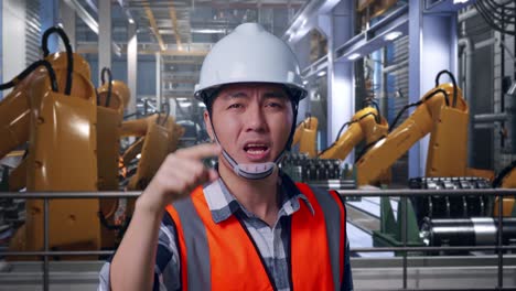 close up of angry asian male engineer with safety helmet shouting at someone while standing with industrial robotic arms working