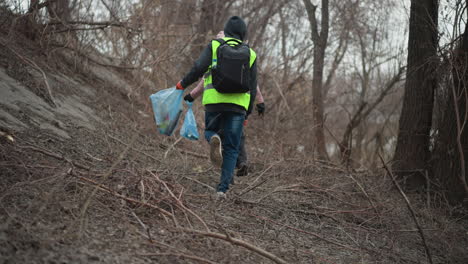 two volunteers wearing reflective vests and gloves walking through forested riverbank area during environmental cleanup, carrying blue plastic bags filled with collected litter to promote nature