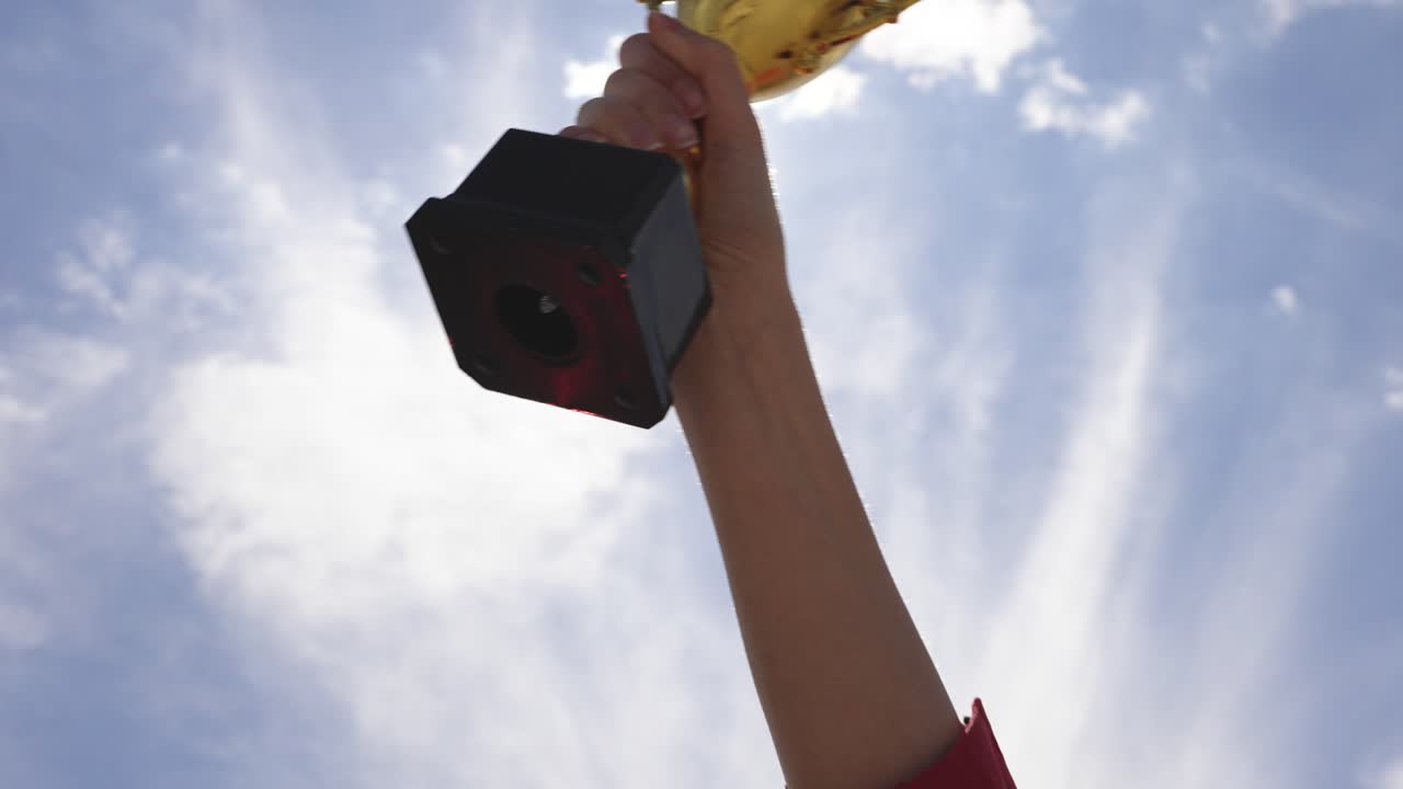 Mano che tiene un trofeo d'oro su sfondo di cielo all'aperto. Concetto di realizzazione, successo, vittoria, motivazione e ricompensa. Rappresenta la vittoria e il raggiungimento degli obiettivi.