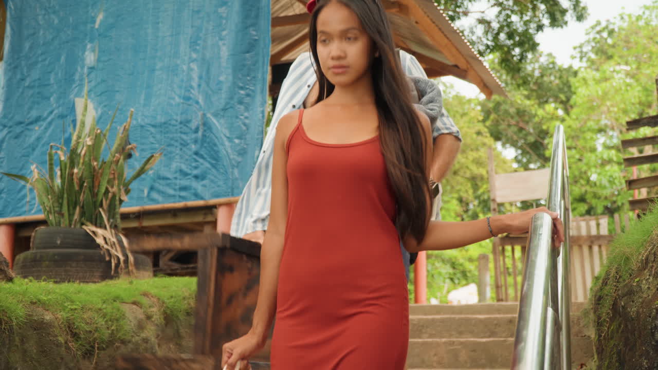 Donna in abito rosso che scende i gradini verso una capanna rustica sulla spiaggia, uomo che la segue con borsa e asciugamano, vegetazione tropicale che incornicia la scena, atmosfera da cerimonia del tè intima, luce dorata calda e passo rilassato.