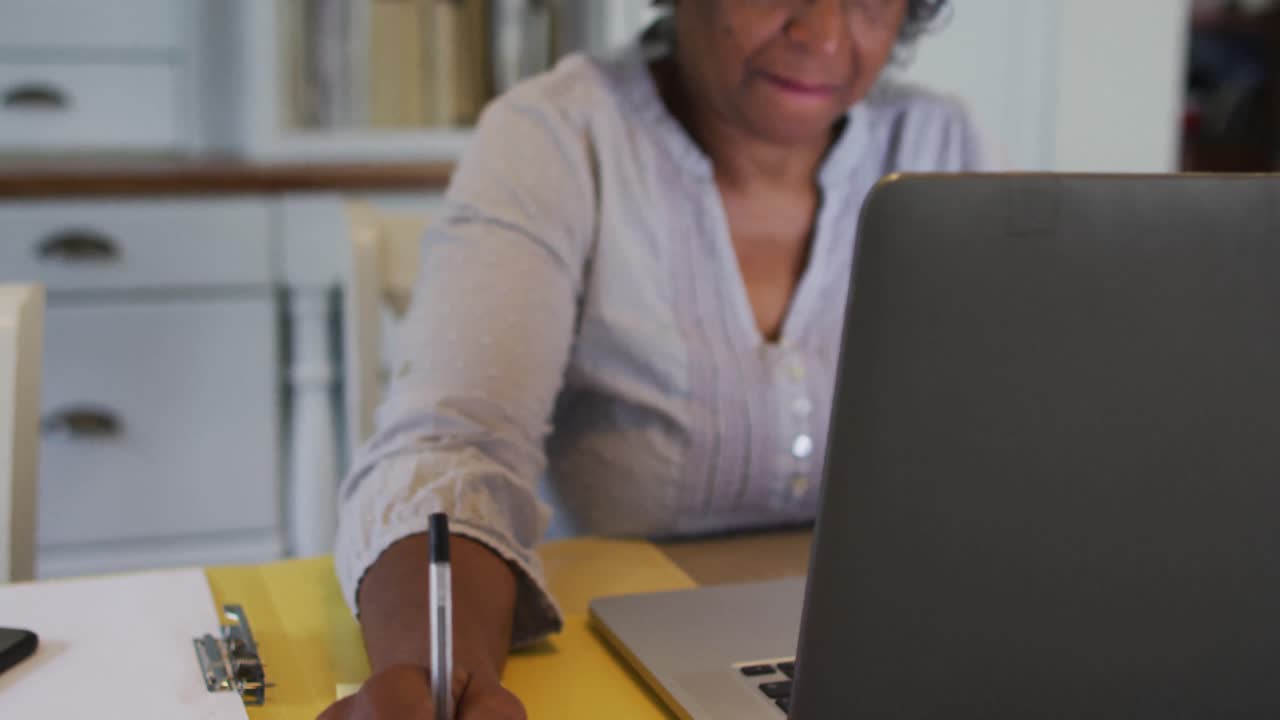 Senior african american woman wearing phone headset taking notes while using laptop at home