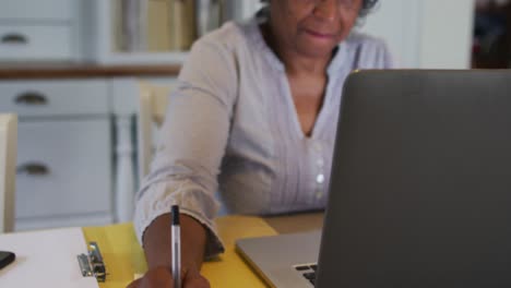 Ältere-Afroamerikanische-Frau-Trägt-Ein-Telefon-Headset-Und-Macht-Sich-Notizen,-Während-Sie-Zu-Hause-Einen-Laptop-Benutzt