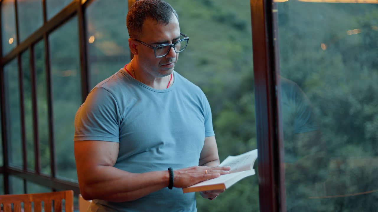 A male person enjoying literature indoors beside window with countryside landscape in the background