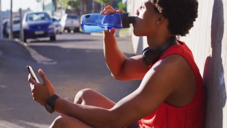 fit african american man exercising in city taking a break, using smartphone, sitting drinking water