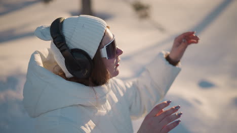 creative spins body while putting on dark specs under warm sunset light wearing white beanie and puffer coat with headphones against blurred snowy forest backdrop evoking energetic winter motion