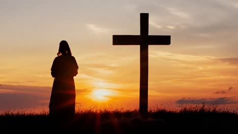 woman stands near a cross, silhouetted against a stunning sunset filled with vibrant orange, red, and yellow hues