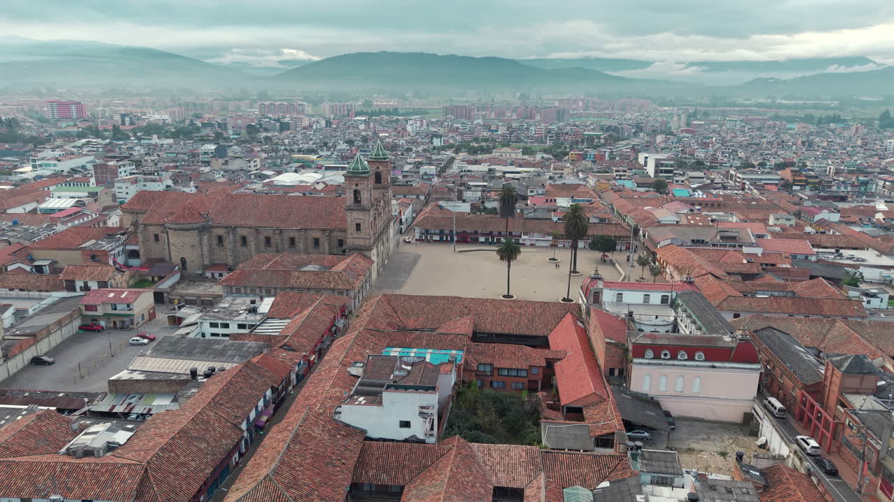 Drone video over Zipaquirá's historic plaza, showcasing red-tiled roofs and classic architecture under a cloudy sky. Captivating wide-angle view with mountain backdrop