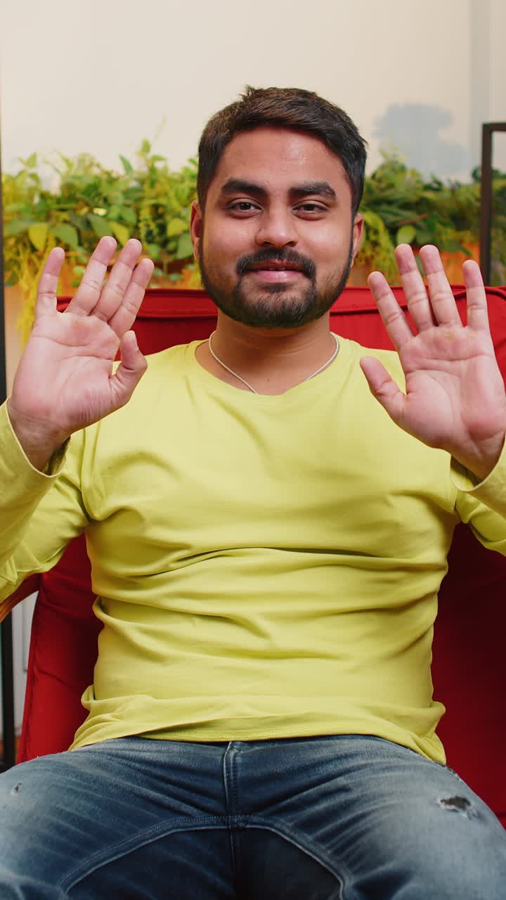 Indian man smiling friendly at camera waving hands gesturing hello hi greeting at home on chair