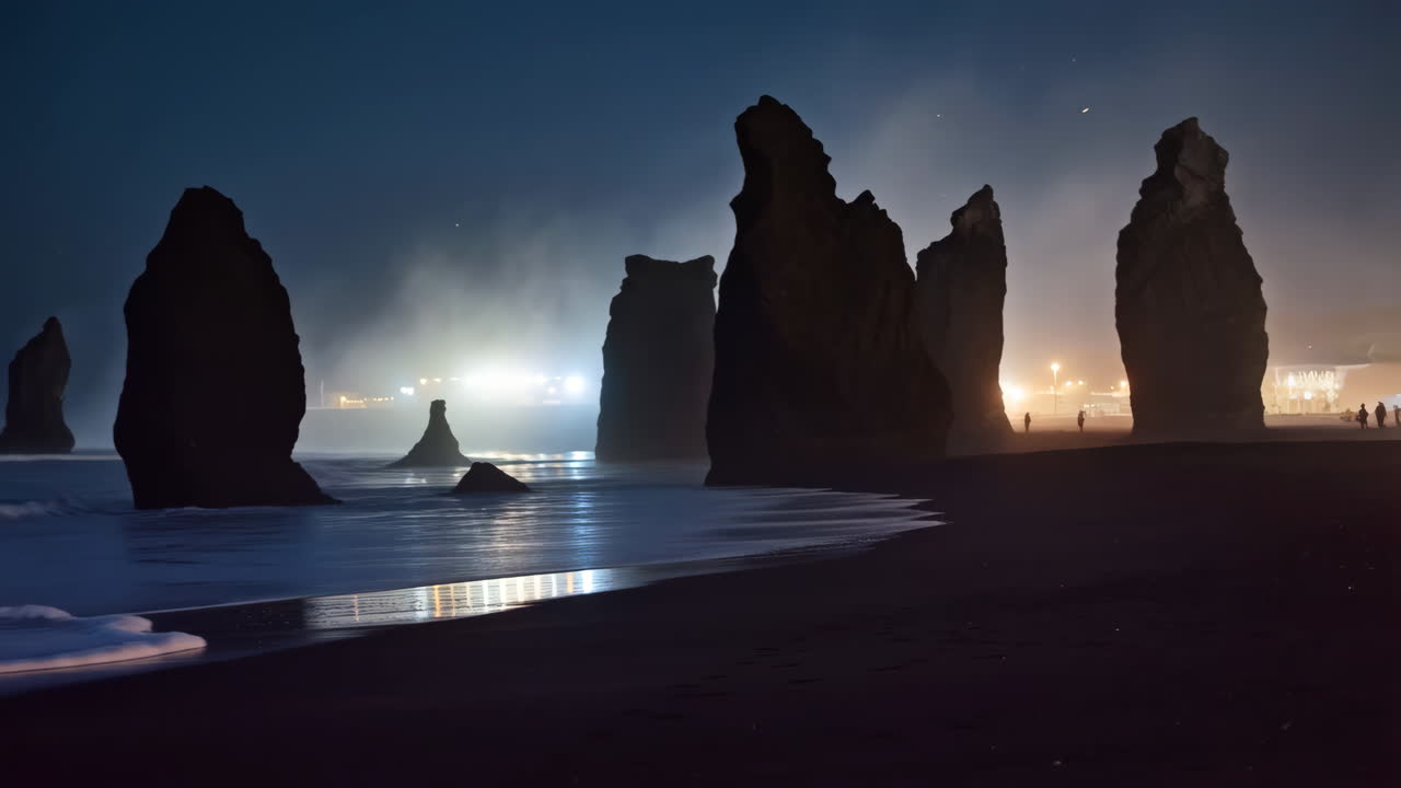 Nighttime view of sea stacks on a dark beach with glowing distant lights and reflections
