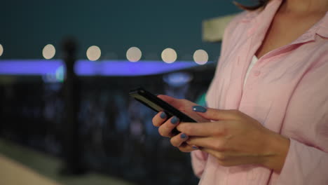 close-up of young woman in pink top holding smartphone, standing by fence with illuminated city lights and blurred bridge in the background at night