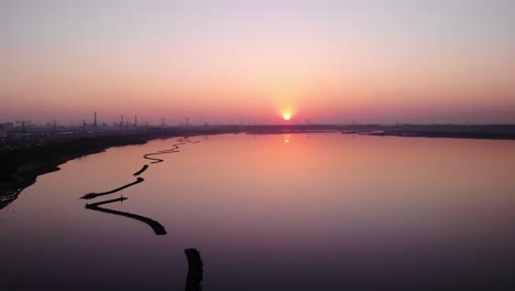 aerial over calm waterway with reflected orange sunset next to silhouetted view of maasvlakte industrial port