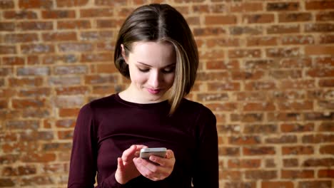 una joven alegre está escribiendo un mensaje en su teléfono inteligente, sonriendo, concepto de comunicación, fondo de ladrillo