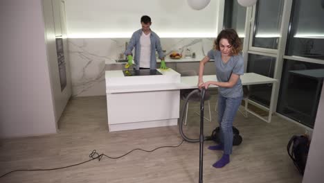 cleaning day. young couple cleaning their apartment - man in gloves wiping up kitchen stove, girl cleaning floor using vacuum cleaner. clean studio kitchen together in the evening. slow motion