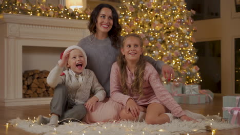 mother, little boy and girl pose and say hello sitting on the floor next to the gifts and the christmas tree