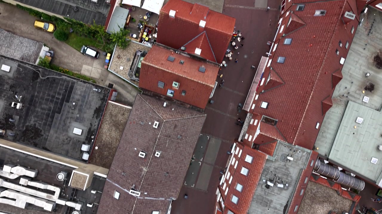Historic red-tile rooftops cluster around cobblestone crossroads in small German town center, parked cars line quiet street beside stone fountain and old brick rowhomes. Aerial drone top down flyover