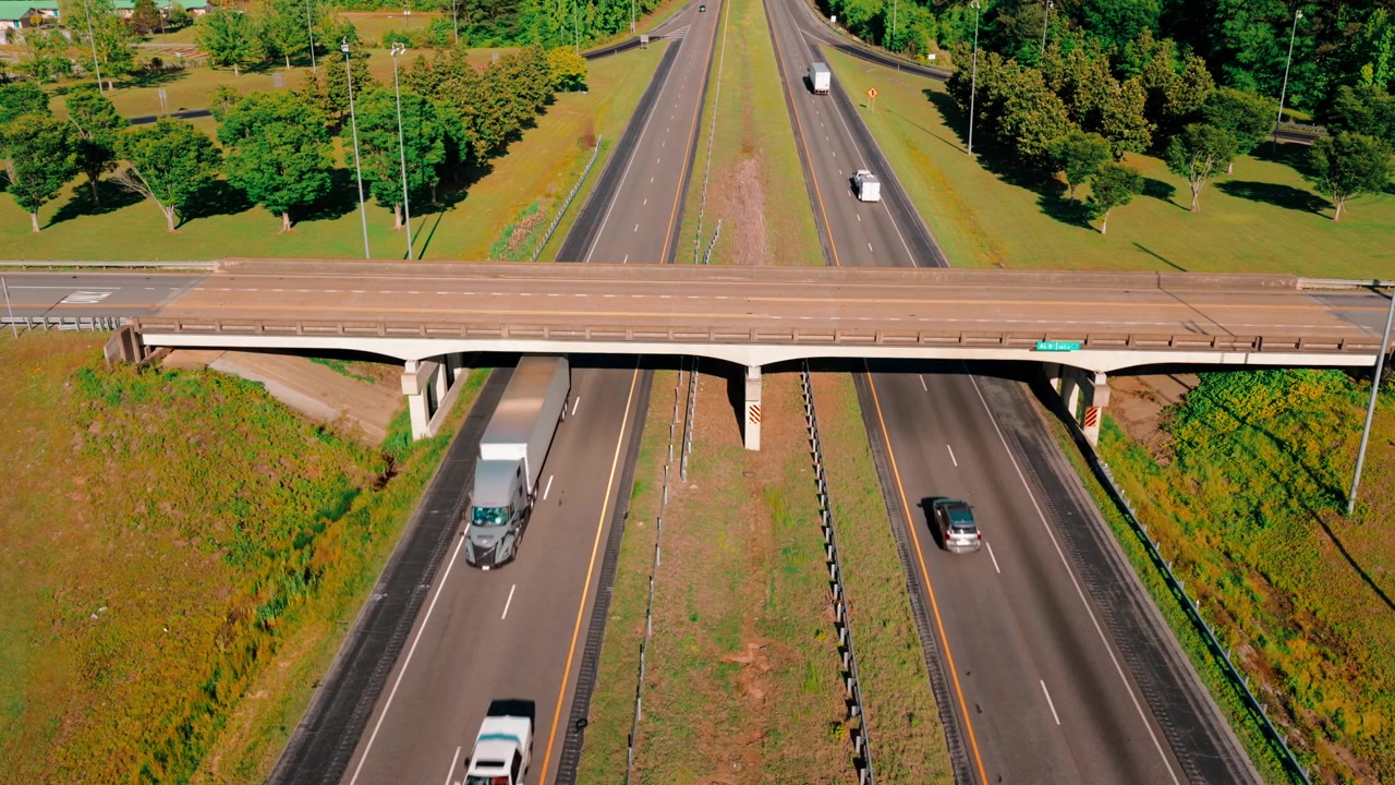 I-20 Aerial Alabama. Symmetrical drone view of a divided highway with a concrete overpass in the foreground, trucks and cars traveling both directions past a wide grassy median and rows of trees.