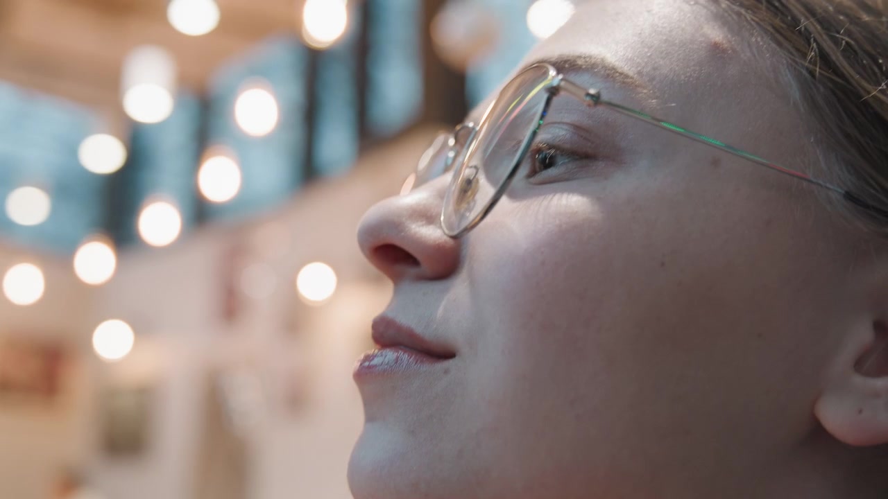 Giovane donna bianca che si sistema gli occhiali e lo chignon in un bistrò con calma curiosità. Le montature delicate catturano caldi riflessi bokeh, le viste di profilo mostrano un'espressione gentile, dettaglio di accessorio moderno, ambiente interno creativo.
