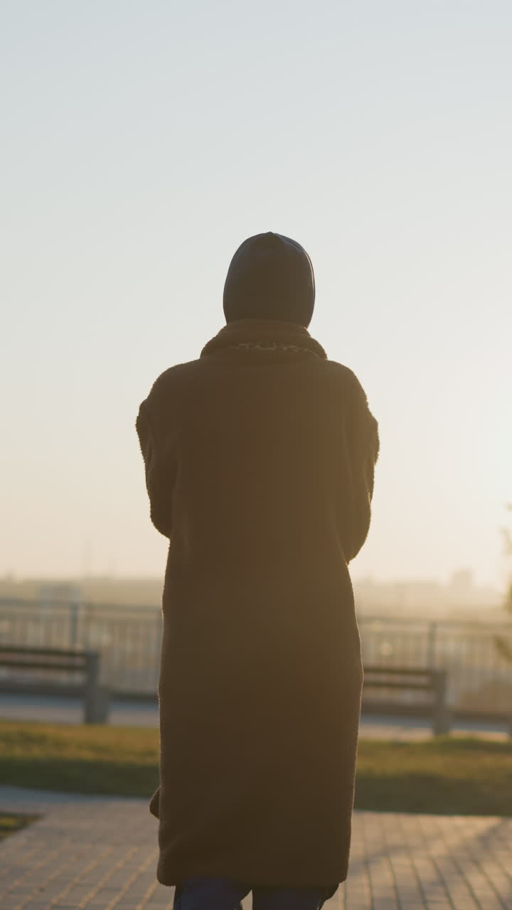 일몰에 공원을 가로질러 천천히 그리고 슬프게 걸어가는 여자의 뒷면 사진. 그녀의 실루은 황금 시간 빛에 의해 부드럽게 조명되어 평온하지만 우울한 분위기를 만니다.