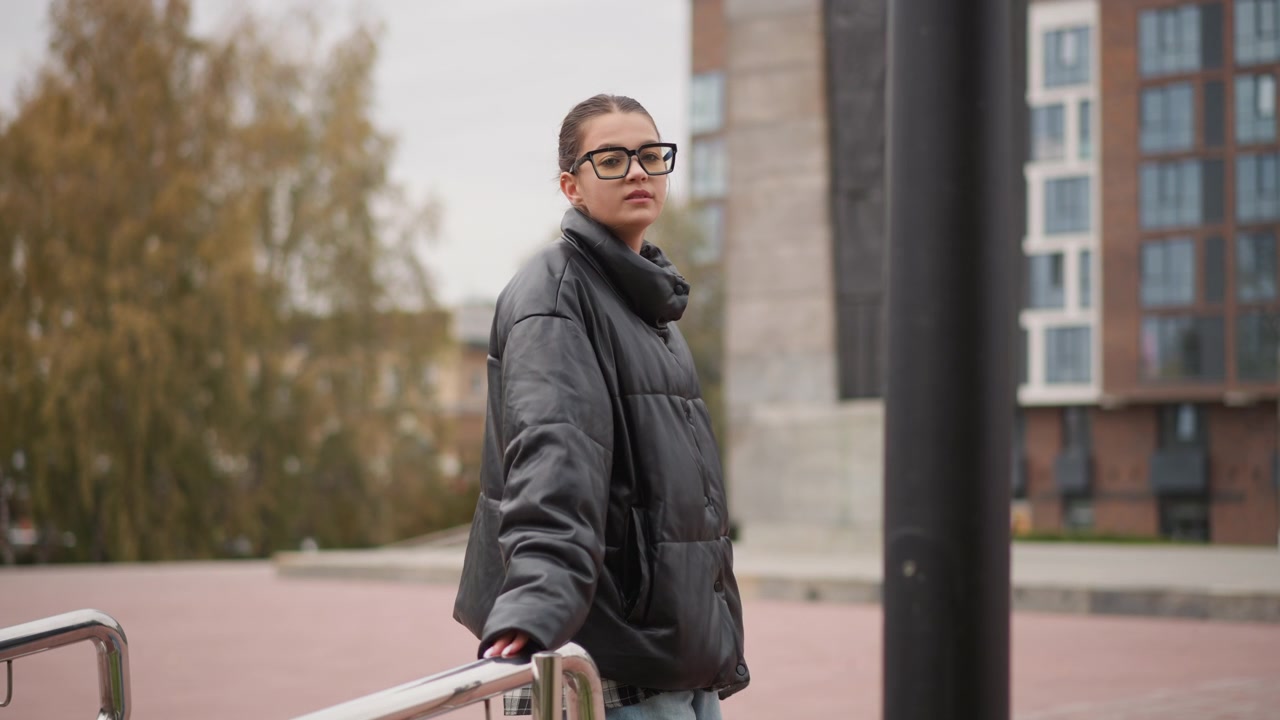 Woman with glasses and bun hairstyle walks slowly along outdoor metal handrail holding it for support. Woman wears black puffer jacket over plaid shirt visible at hemline in front of urban