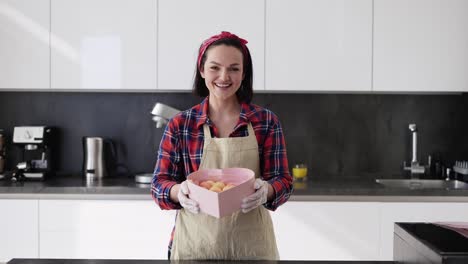 Mujer-Sonriente-En-Delantal-Presenta-Una-Caja-Con-Galletas-De-Macarrones-En-La-Cocina