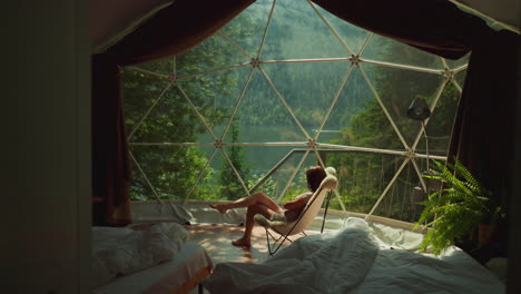 resting female enjoys breathtaking view from modern bedroom window. rented glamping house in forest near lake. atmospheric place to relax and enjoy beauty of nature