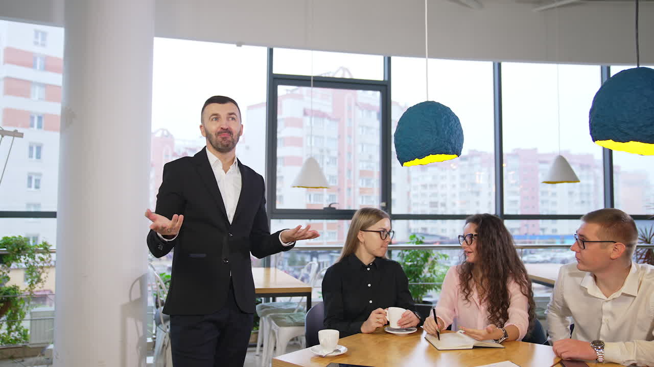 Mid-aged neat businessman in suit stands at the desk and talks to the camera. People sitting at the table having a talk among each other.