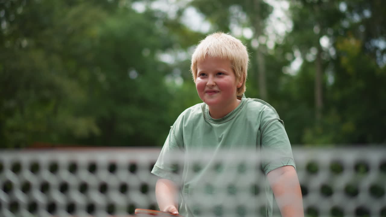 Menino branco jogando tênis de mesa com raquete e rede, sorrindo e segurando a raquete em quadra ao ar livre, momento esportivo jovial com plano de fundo de árvores verdes, expressão brincalhona focada e vibração recreativa
