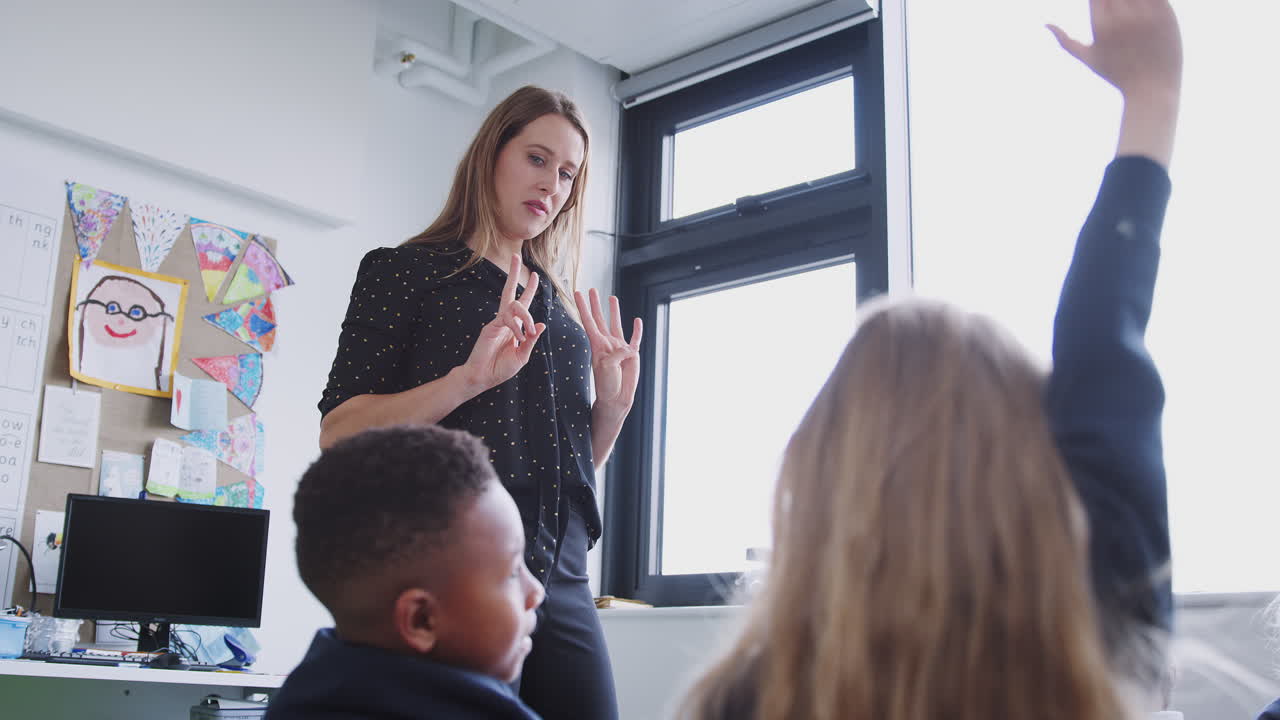 Female primary teacher standing at the front of primary school class, kids raising hands, low angle