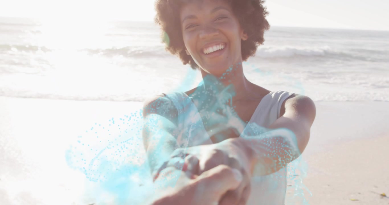 Femme tenant la main de son partenaire, créant un effet de particules bleues tourbillonnantes autour de leurs bras sur une plage, démo technologique.