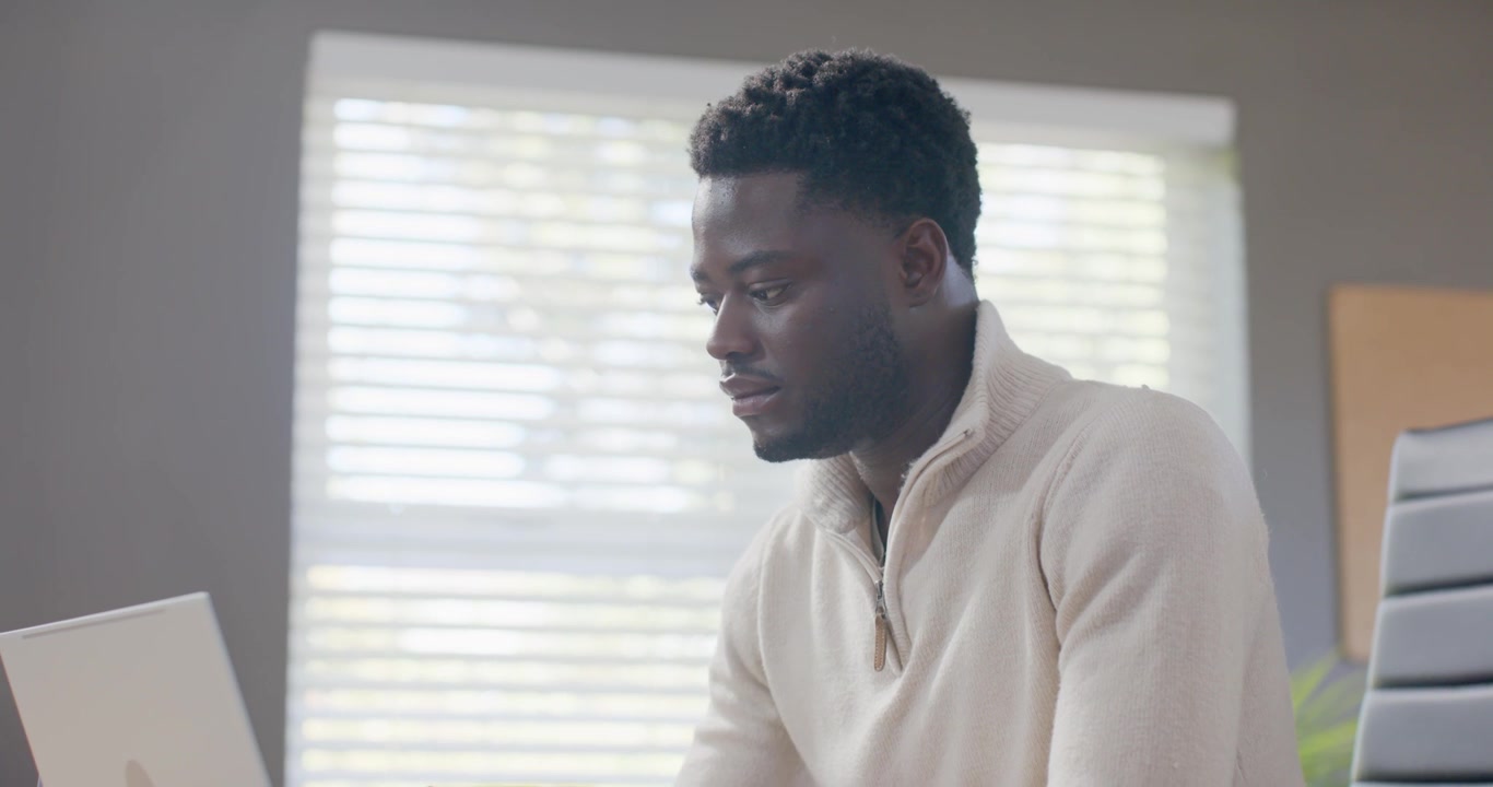 African American man in sweater at desk by blinds holding mug reading laptop then typing replies