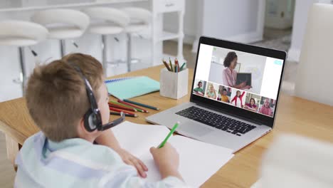 Schoolboy-using-laptop-for-online-lesson-at-home,-with-diverse-teacher-and-class-on-screen