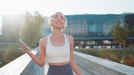 happy young woman jogger listening to music via headphones and smartphone walking in city park